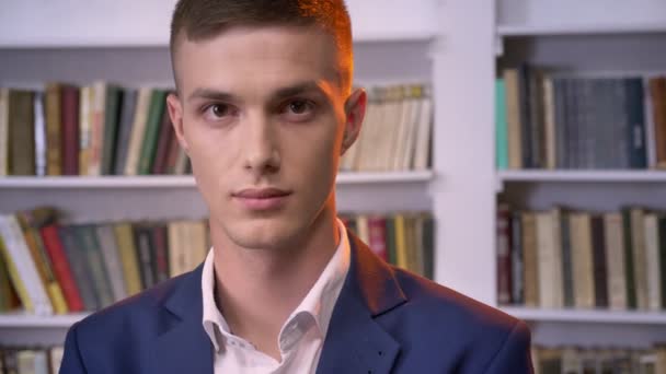 Jeune homme d'affaires attrayant regarde à la caméra, souriant, bibliothèque sur fond — Video