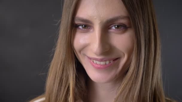 Ansikte av ung blyg flicka tittar på kameran, leende, grå bakgrund — Stockvideo