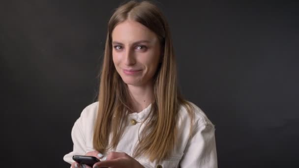 Молодая крутая девушка шлет сообщение на смартфоне, смотрит на камеру, коммуникационная концепция, серый фон — стоковое видео