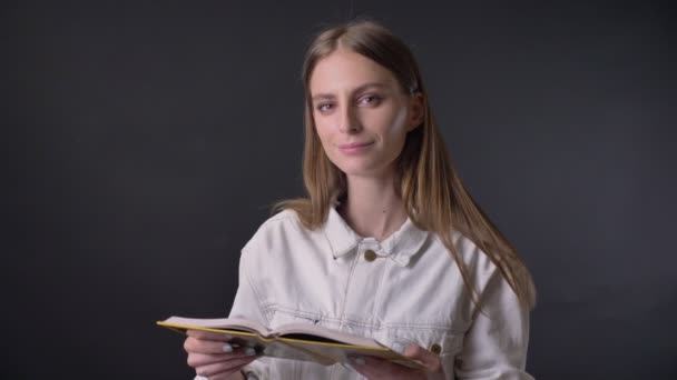 Ung söt tjej läser bok, titta på kameran, leende, grå bakgrund — Stockvideo