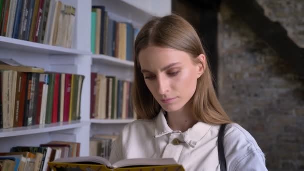 Joven chica seria está leyendo libro, biblioteca en el fondo — Vídeo de stock