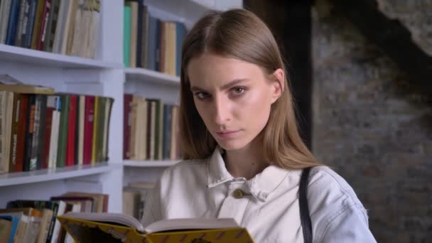 Σοβαρή κοπέλα διαβάζει το βιβλίο, βλέποντας στο κάμερα, βιβλιοθήκη στο φόντο — Αρχείο Βίντεο