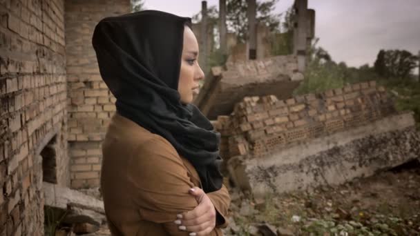 Mujer musulmana joven en hijab de pie cerca de edificio en ruinas y mirando a la cámara con expresión asustada y preocupada, ruina en el fondo — Vídeos de Stock