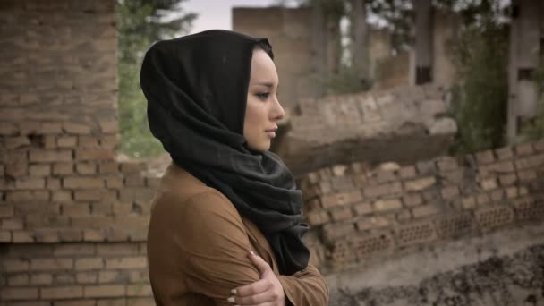 Young refugee muslim woman in hijab standing near ruined building and looking at camera with frightened and scared expression during war — Stock Video