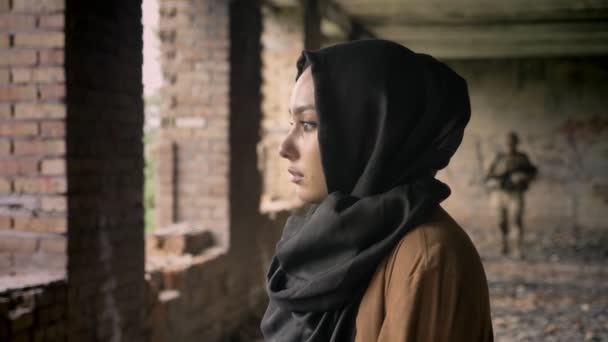 junge traurige Muslimin im Hidschab stehend, Soldat mit Munition nähert sich Frau, verlassenes Gebäude, Terrorismus