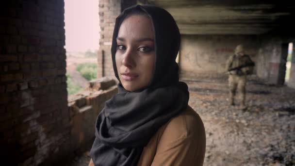 junge muslimische Frau im Hidschab, weinend und in die Kamera blickend, Soldat mit Waffe im Hintergrund, verlassenes Gebäude, Terrorkonzept
