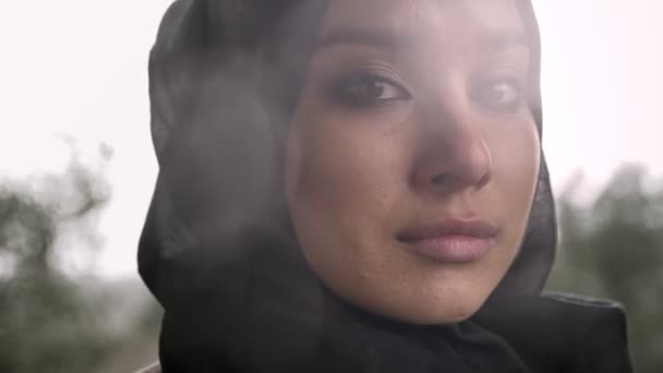 Retrato de jovem mulher muçulmana triste no hijab olhando para a câmera e chorando, tempo chuvoso no fundo — Vídeo de Stock
