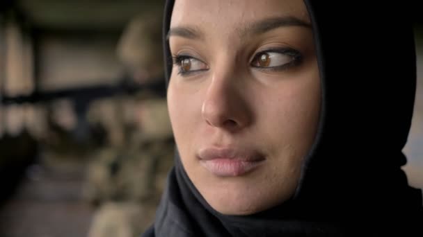 Близький портрет молодої впевненої мусульманки, що повертається і дивиться на камеру, озброєного солдата на задньому плані, концепція війни — стокове відео