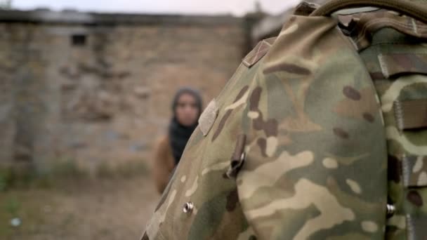 Bewaffneter Soldat geht neben stehender muslimischer Frau im Hidschab, Frau blickt in Kamera, bewegliche Kamera, Backsteinruine Hintergrund