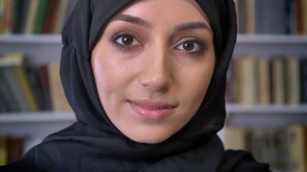 Молода красива мусульманська дівчина в хіджабі дивиться вниз в книгу, дивиться на камеру, релігійну концепцію, книжкову полицю на фоні, розмитий фон — стокове відео