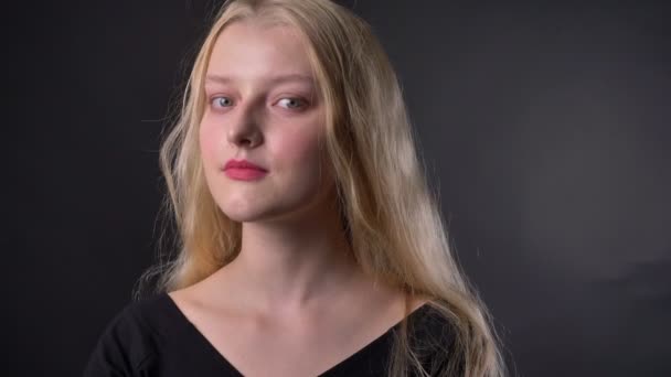 Jeune belle fille blonde fatiguée avec du rouge à lèvres rose regarde à la caméra, cheveux touchants, fond gris — Video