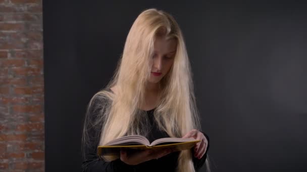 Jeune fille blonde mignonne avec du rouge à lèvres rose lit le livre, regardant à la caméra, souriant, fond gris — Video