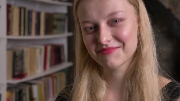 Joven chica rubia linda con lápiz labial rosa está mirando a la cámara, sonriendo, biblioteca en el fondo, fondo borroso — Vídeos de Stock