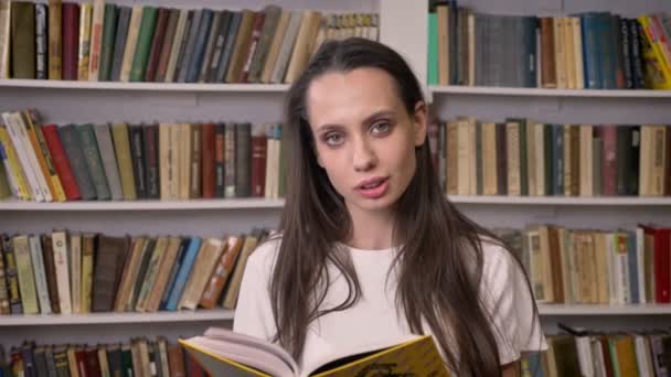 Молодая сексуальная брюнетка читает книгу в библиотеке, смотрит в камеру, улыбается — стоковое видео
