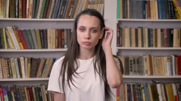 Молодая сексуальная брюнетка стоит в библиотеке и подправляет волосы. — стоковое видео