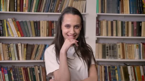 Jovem menina morena feliz está de pé na biblioteca, rindo — Vídeo de Stock