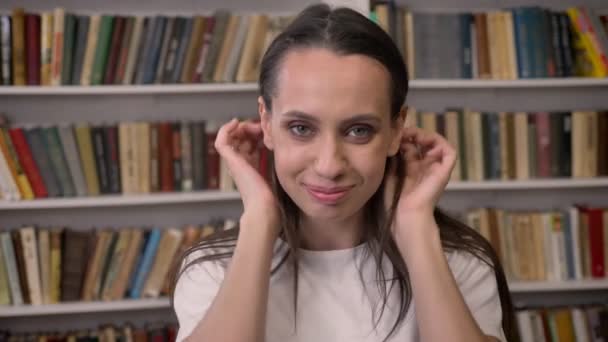 Joven morena feliz chica está de pie en la biblioteca, metiendo el pelo, riendo — Vídeos de Stock