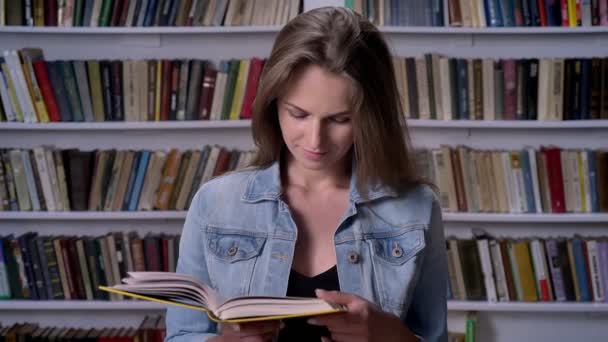 Joven linda mujer está leyendo libro en la biblioteca, mirando a cámara, riendo, estante en backgorund — Vídeo de stock