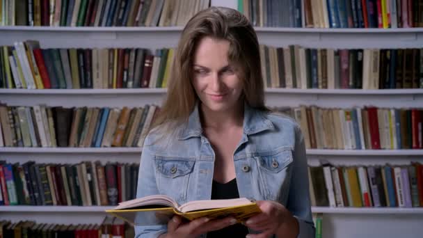 Молодая симпатичная женщина читает книгу в библиотеке, смотрит на камеру, улыбается, книжится на заднем плане — стоковое видео