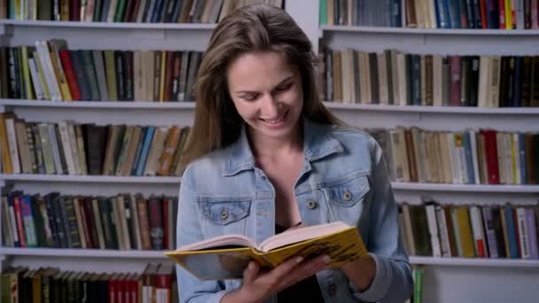 Jovem mulher bonita está lendo livro na biblioteca, sorrindo, estante em backgorund — Vídeo de Stock