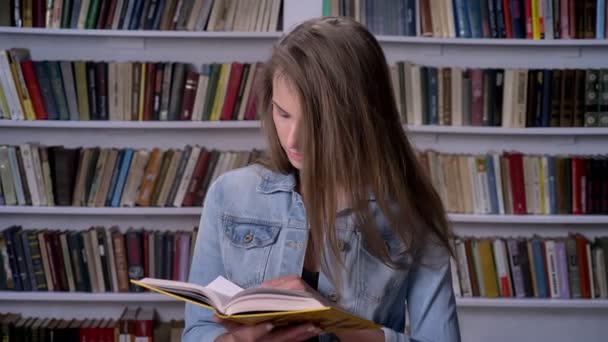 Молодая серьезная женщина читает книгу в библиотеке, смотрит в камеру, книжная полка на бэкгорунде — стоковое видео