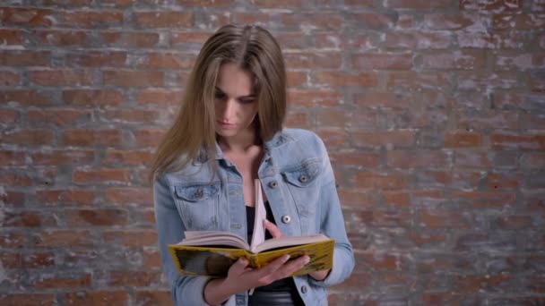 Jovem bonito mulher está lendo livro, assistindo a câmera, sorrindo, tijolo backgorund — Vídeo de Stock