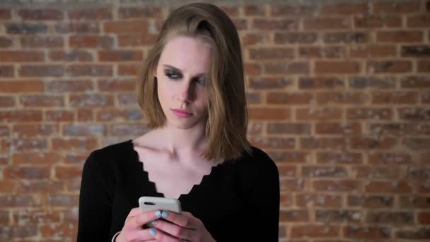 Fiatal szexi lány, füstös szemek gépelés üzenet a smartphone, gondolkodási folyamat, a kommunikáció fogalma tégla háttér — Stock videók