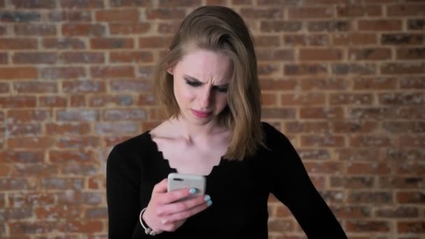 Ung sexig tjej med sotade ögon tittar på foton på smartphone, missnöje, kommunikationskoncept, tegel bakgrund — Stockvideo