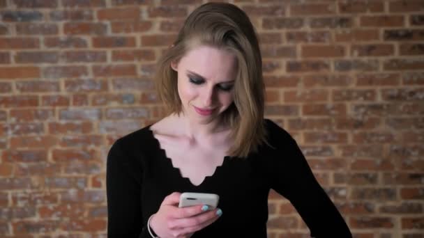 Menina sexy nova com olhos fumegantes está assistindo fotos no smartphone, rindo, conceito de comunicação, fundo de tijolo — Vídeo de Stock