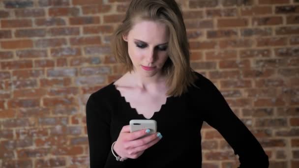 Dumanlı gözler ile genç seksi kız fotoğrafları smartphone, kıkır kıkır gülme, iletişim kavramı, duygu kavramı, tuğla arka plan üzerinde izliyor — Stok video