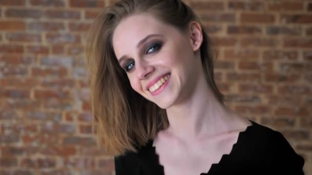 Jeune fille charmante avec des yeux fumés regarde à la caméra, souriant, concept flirt, fond de brique — Video