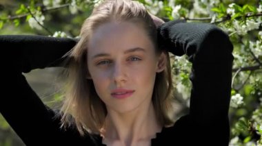 Genç çekici kız saç parkta gündüz yaz, kameraya izlerken, bloom ağaç arka plan üzerinde dokunuyor