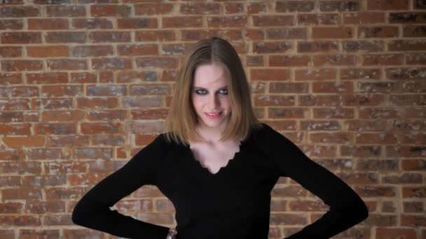 Young strong girl with smoky eyes is standing with hands on hips, staring at camera, laughing, brick background — Stock Video