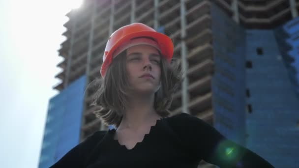 Young proud girl builder with hemlet is standing with hands on hips in daytime in summer, building concept, urban concept, building on background, bottom view — Stock Video