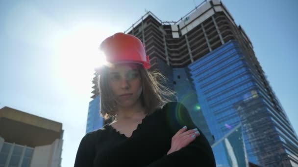 Молодая сильная девушка строитель с камушком стоит со скрещенными руками в дневное время летом, строительная концепция, городская концепция, здание на заднем плане, вид снизу — стоковое видео