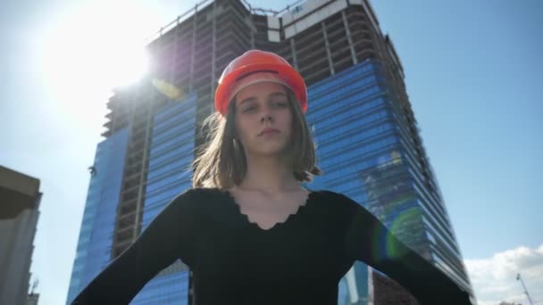 Молода впевнена дівчина-будівельник з клейнодом стоїть з руками на стегнах вдень влітку, повертаючи голову, будуючи концепцію, міську концепцію, будуючи на фоні — стокове відео