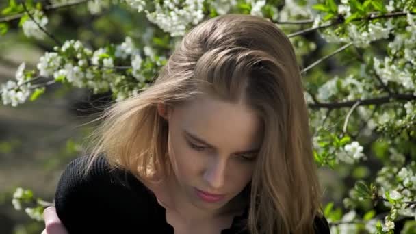 Chica de la liana joven se está abrazando en el parque durante el día en verano, mirando a la cámara, árbol de flores en el fondo — Vídeo de stock