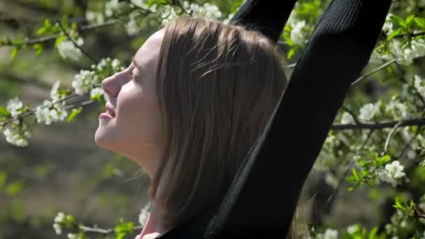 Joven chica inspirada se estira en el parque durante el día en verano, mirando a la cámara, árbol de flores en el fondo — Vídeos de Stock