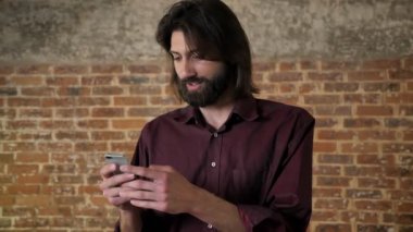 Sakallı genç güzel esmer adam akıllı telefon, gülüyor, tuğla arka plan, iletişim kavramı fotoğrafları izlerken
