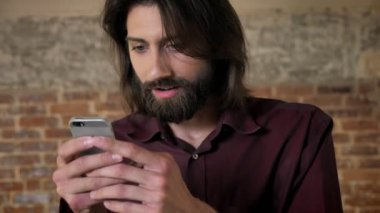 Sakallı genç şok esmer adam akıllı telefon, tuğla arka plan, iletişim kavramı fotoğraflar izliyor