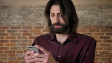 Sakallı genç çekici esmer adam akıllı telefon, kamera, tuğla arka plan, iletişim kavramı, izlerken izliyor