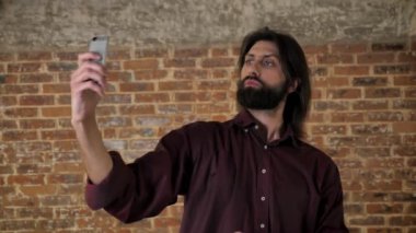 Sakallı genç çekici esmer adam akıllı telefon, dalga geçmeyi, gülüyor, tuğla arka plan, iletişim kavramı üzerinde video yapıyor