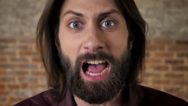 Mladá bruneta muž s vousy se dívá na kameru, křičí, cihlové pozadí — Stock video