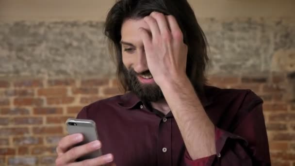 Joven morena atractiva hombre con barba está viendo en el teléfono inteligente, metiendo el pelo, fondo de ladrillo, concepto de comunicación — Vídeos de Stock