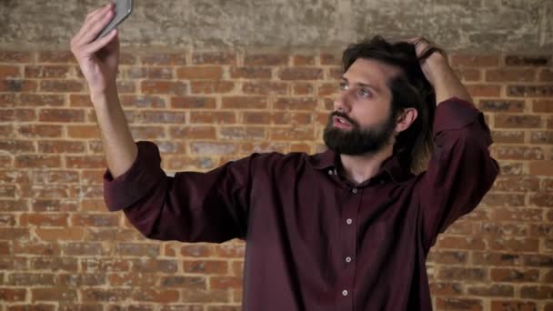 Young attractive brunette man with beard is making selfie on smartphone, touching hair, brick background, communication concept — Stock Video