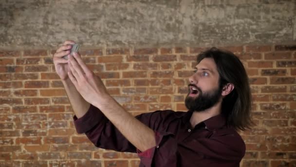 Young attractive brunette man with beard is making video on smartphone, fooling around, brick background, communication concept — Stock Video