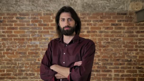 Young confident brunette man with beard is watching at camera, arms are crossed, brick background — Stock Video