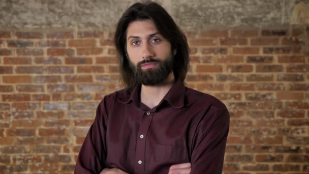 Young confident brunette man with beard is watching at camera, arms are crossed, smiling, brick background — Stock Video