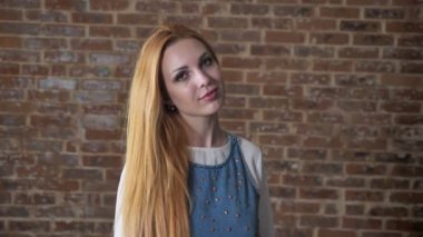 Genç çekici sarışın kız kameraya, dokunmadan saç, flört kavramı, tuğla arka plan izliyor
