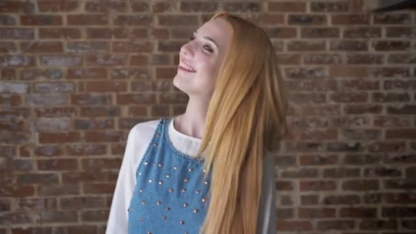 若い魅力的なブロンドの女の子は髪の毛で遊んでカメラ、浮気の概念、レンガの背景を見て — ストック動画